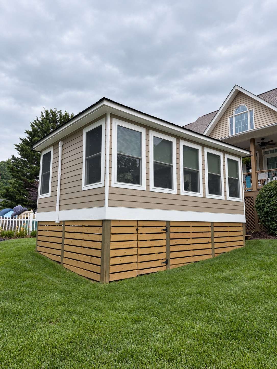 Finished tan lap siding with white trim and a wood-slat skirt on a sunroom addition